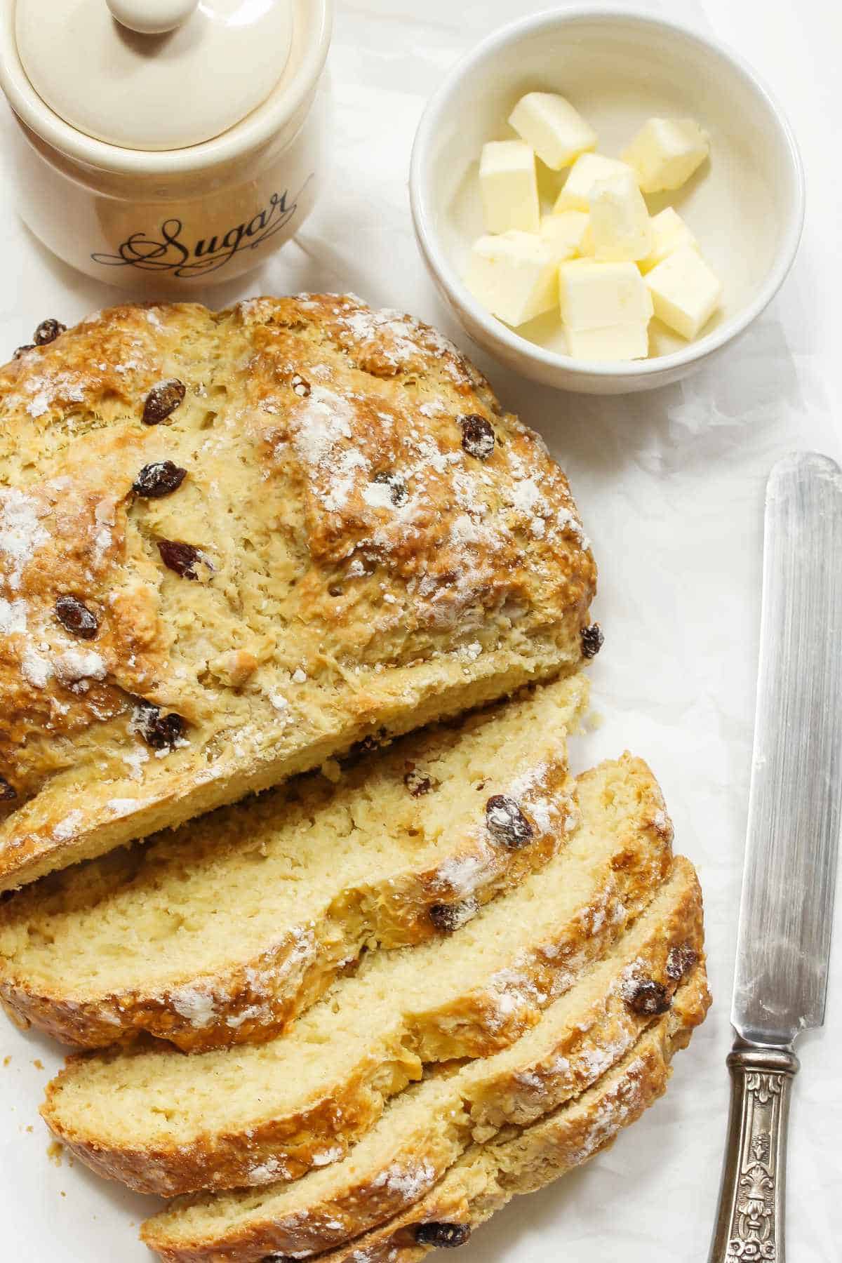 Soda bread with raisins sliced on a table with cubes of butter and sugar.