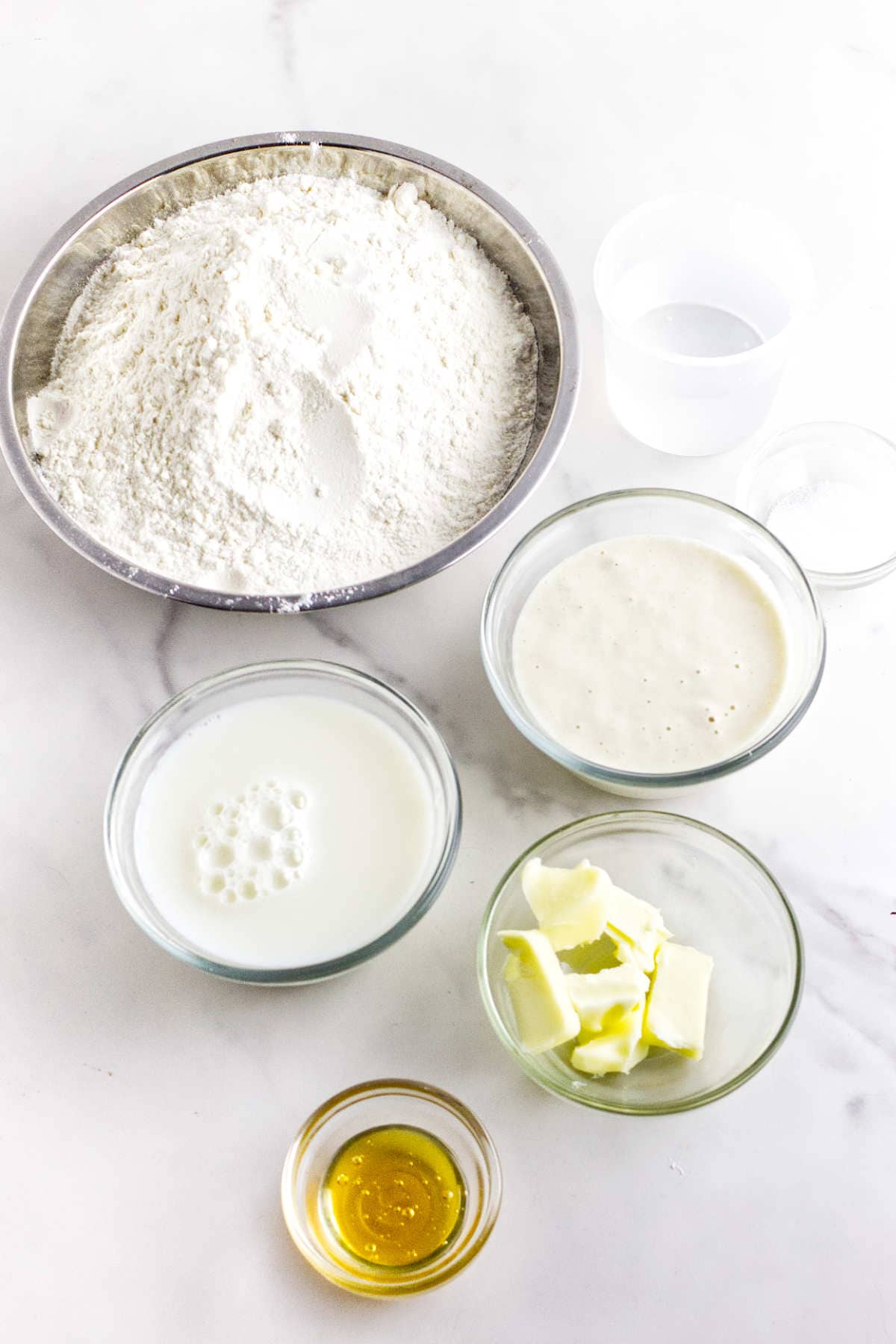 A collection of baking ingredients is displayed on a white surface, including a bowl of flour, a glass of water, bowls of milk and creamy liquid, chunks of butter, and a small bowl of honey.