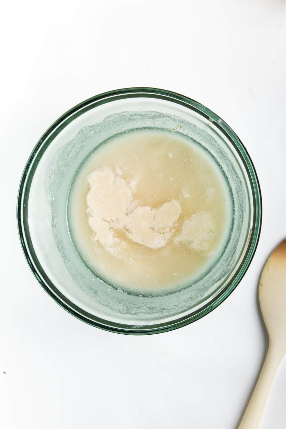 A glass bowl containing a yeast mixture is placed on a white background. A wooden spoon lies beside the bowl. The yeast appears partially dissolved in liquid.