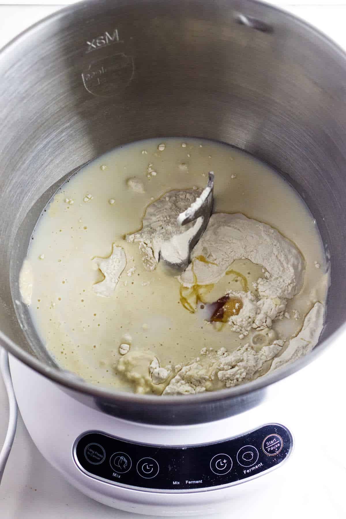 A mixing bowl with flour, milk, and a small amount of liquid ingredients on top of an induction cooktop. The cooktop has a digital control panel with buttons and a small display screen. The scene appears ready for mixing or preparation.