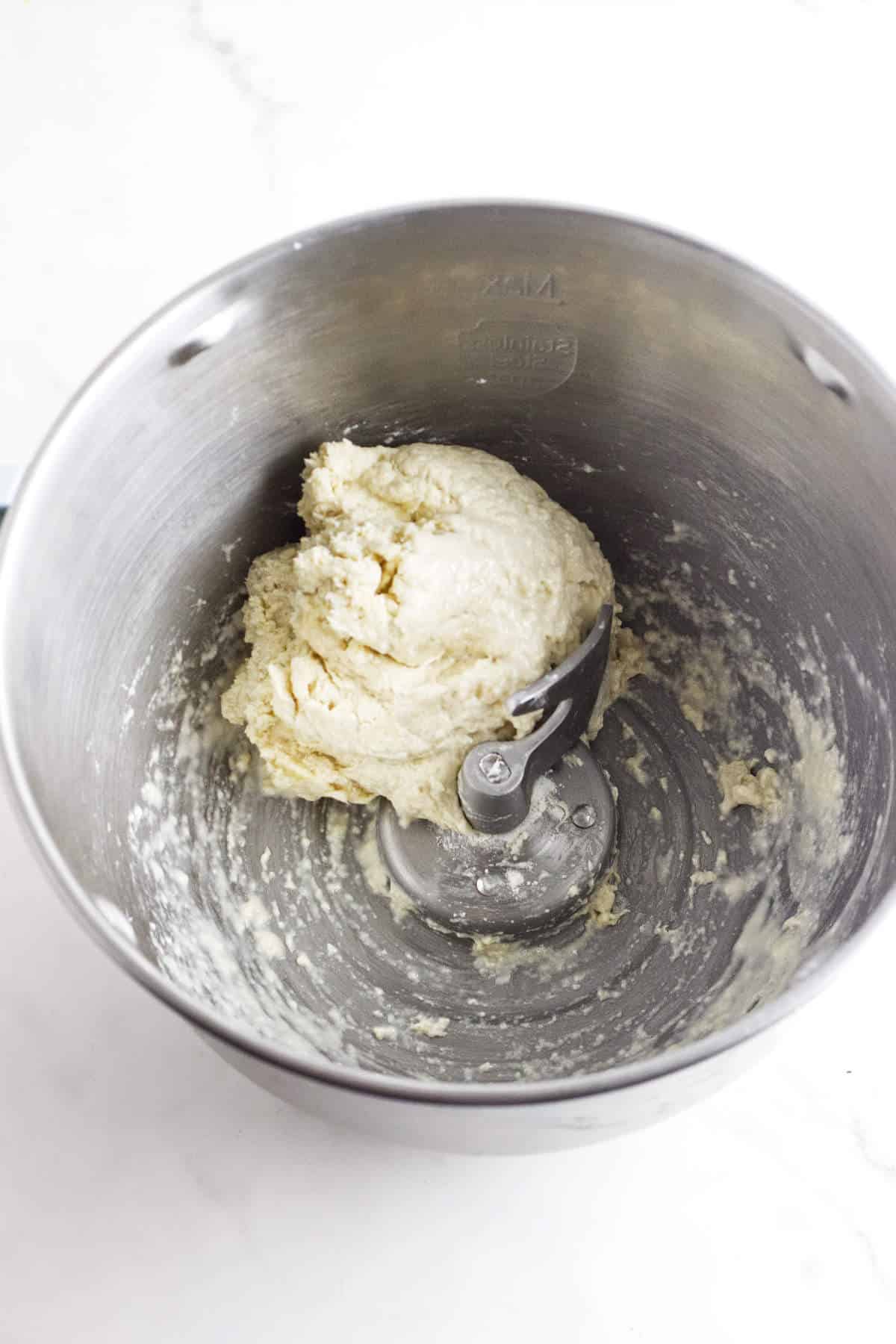 A metal mixing bowl containing a ball of dough, partially mixed with a dough hook attached. The sides of the bowl have traces of flour and dough, indicating recent mixing. The setup is on a white countertop.