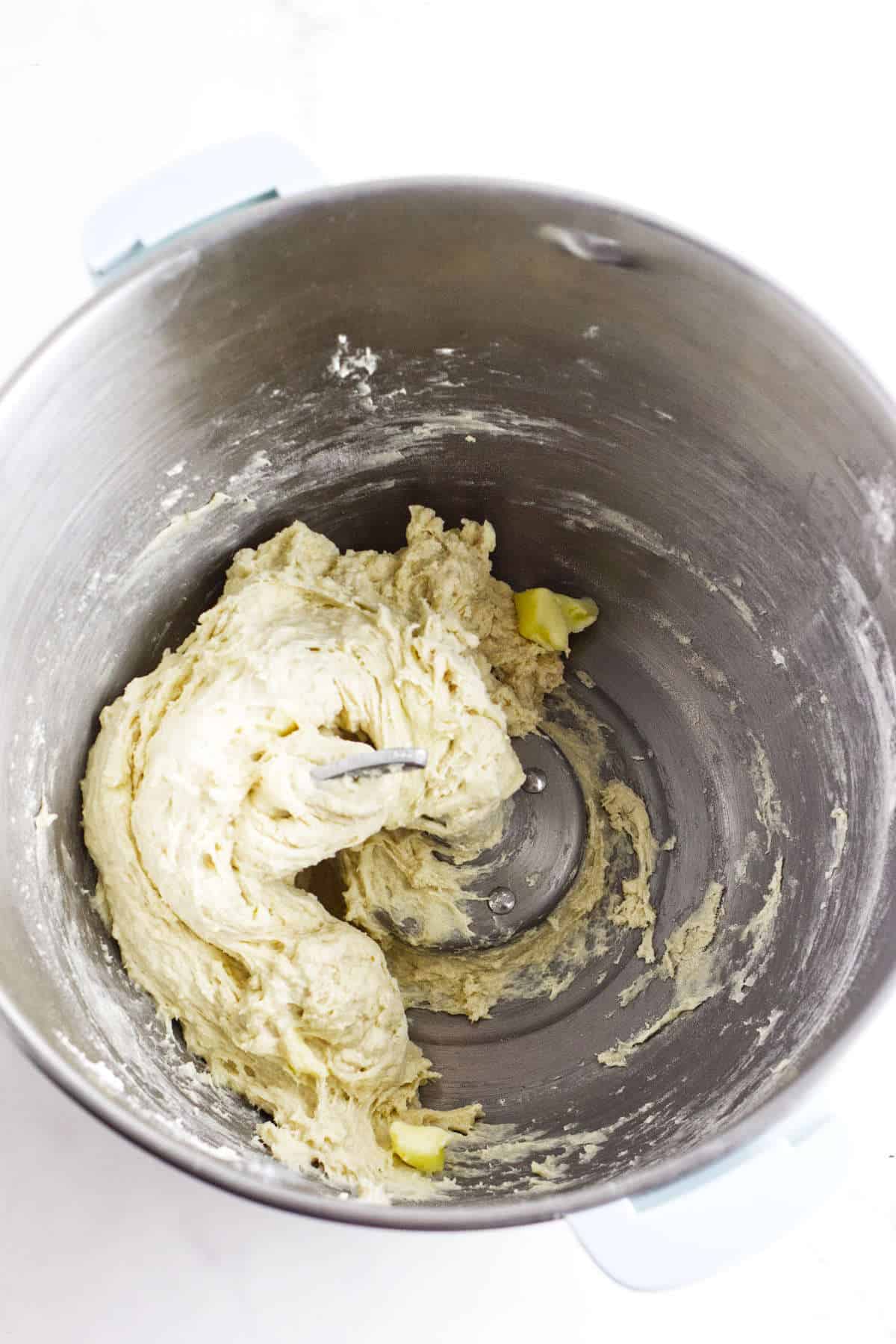 A mixing bowl with partially kneaded dough, with visible chunks of butter, sits on a white countertop. The dough is light in color and appears to be in the early stages of mixing.