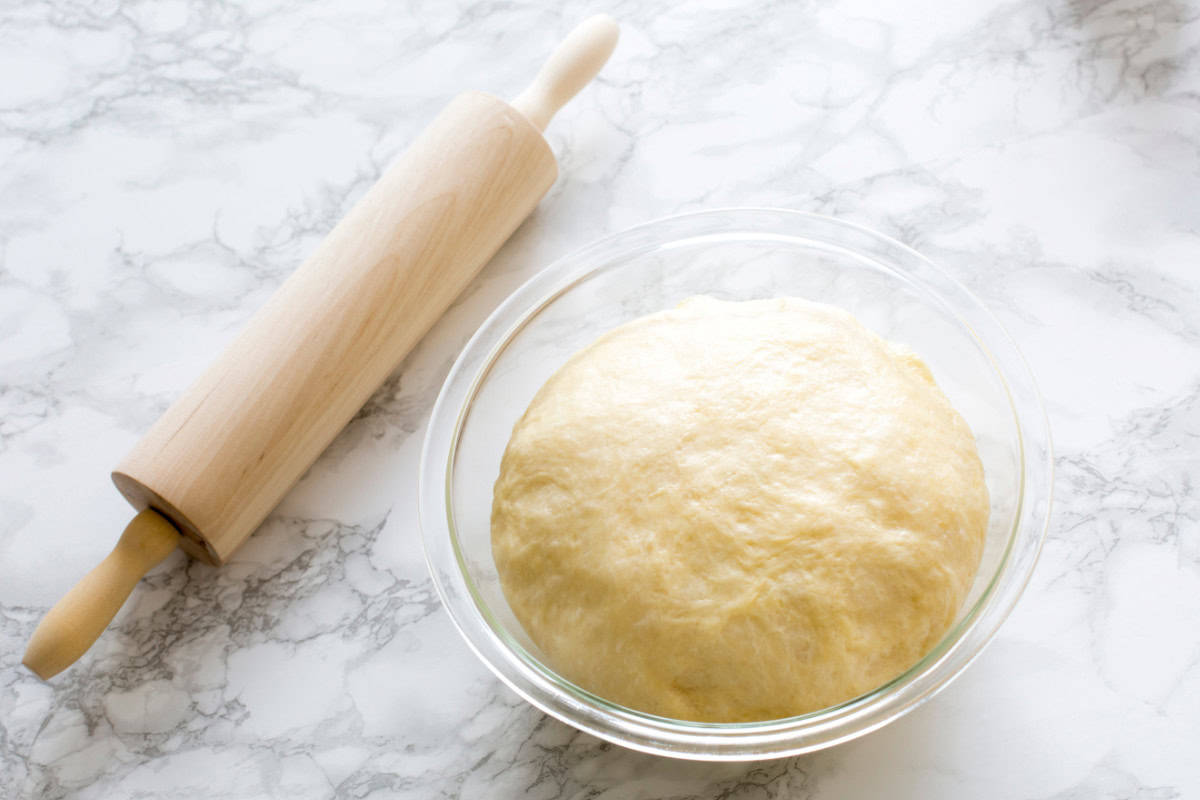 A ball of dough in a glass bowl on a marble surface, proofing slowly, next to a wooden rolling pin.