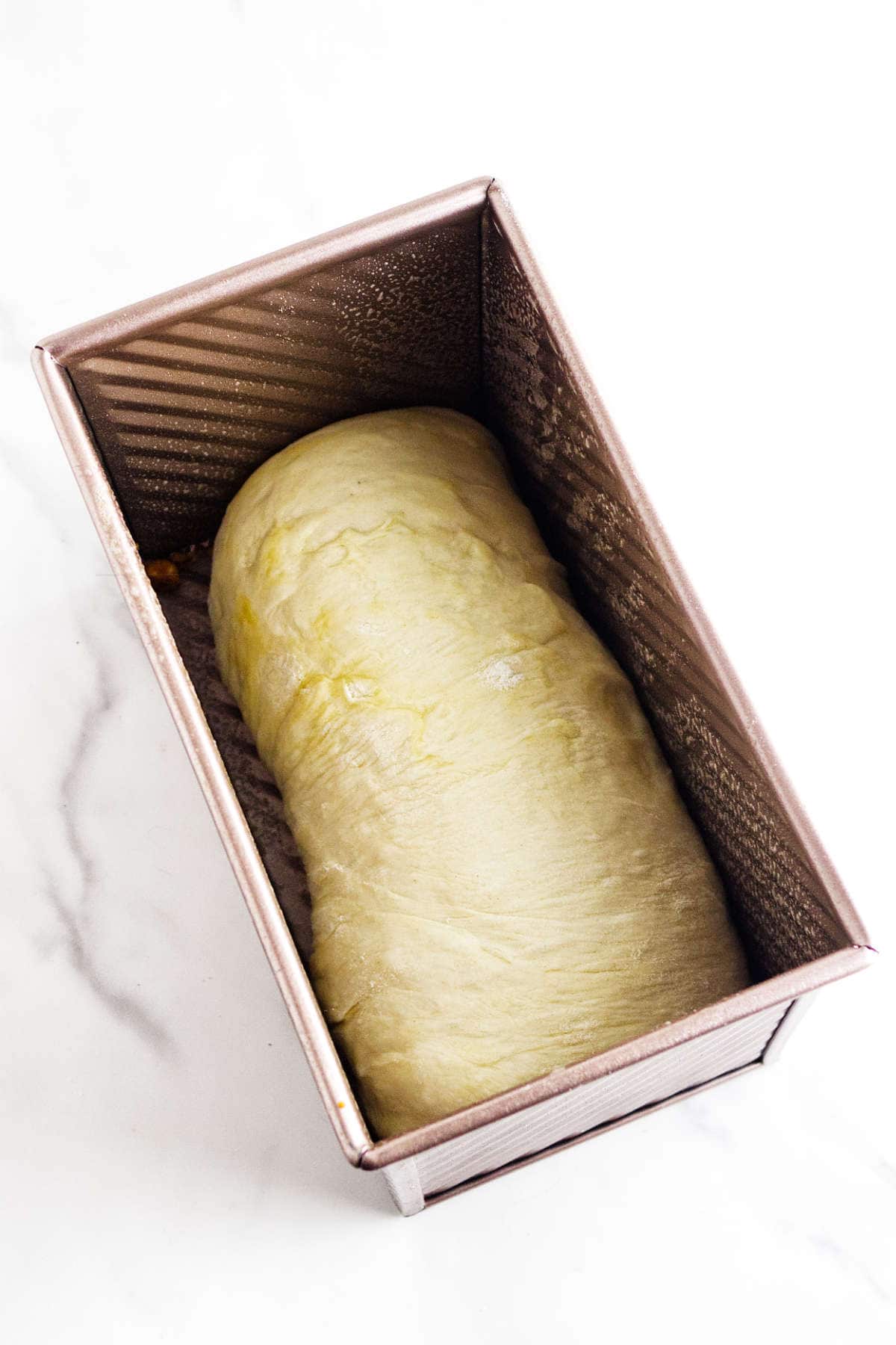 A loaf of bread dough in a rectangular metal pan, ready for baking. The dough has a smooth surface and is lightly brushed with oil. The pan sits on a white marble surface.