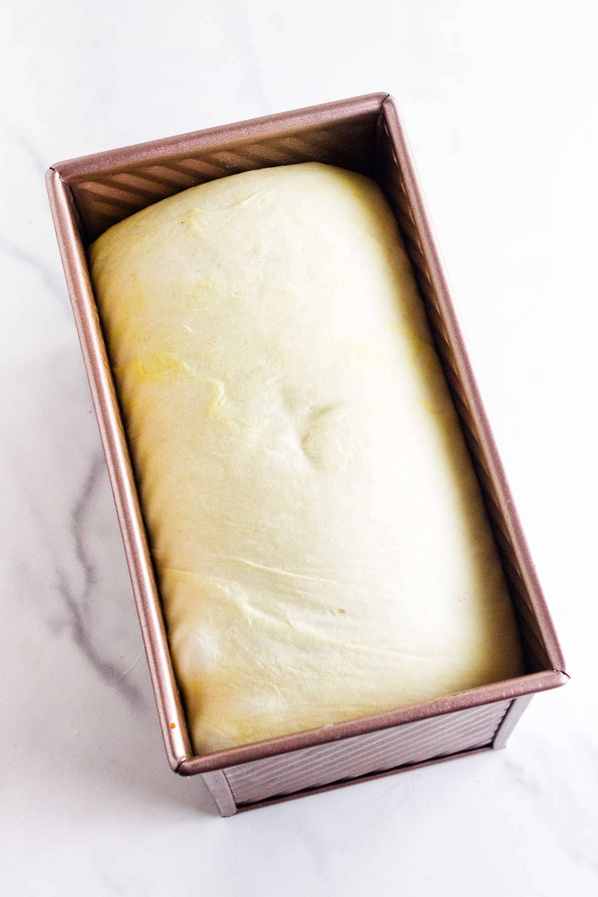 A loaf of bread dough rests in a rectangular baking pan on a marble surface, ready to be baked.