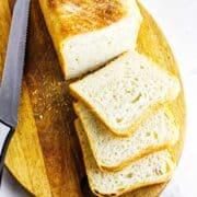 Pain de Mie Pullman bread loaf on a wooden cutting board. Three slices are fanned out in front of the loaf. A serrated knife rests beside the bread. The crust is golden brown and the inside is soft and fluffy.