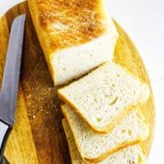 A loaf of Pullman bread on a wooden cutting board with three slices cut. A serrated knife lies beside the bread. The bread has a golden-brown crust and a soft, airy interior, set against a white marble background.