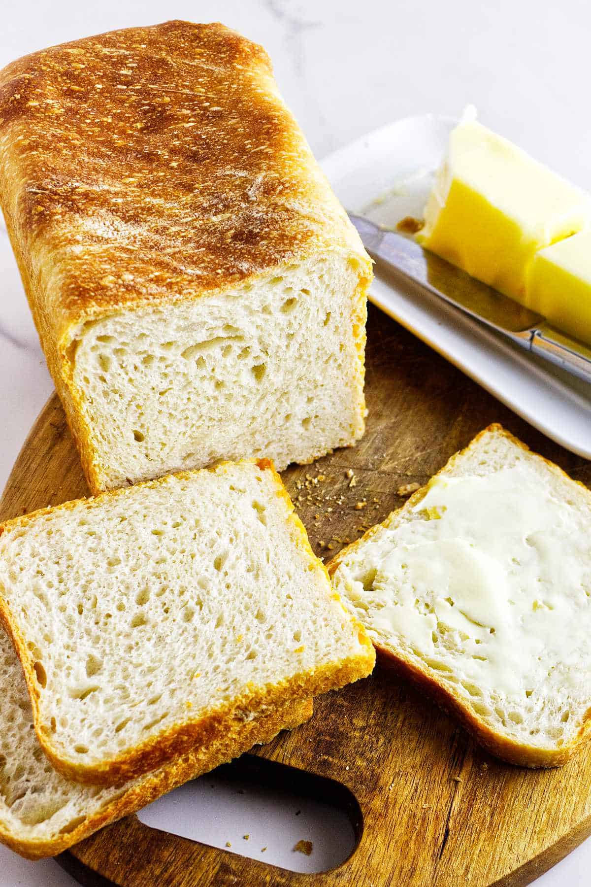A freshly baked loaf of Pain de Mie on a wooden cutting board with two slices cut. One slice is buttered, and a butter dish with a stick of butter is in the background.