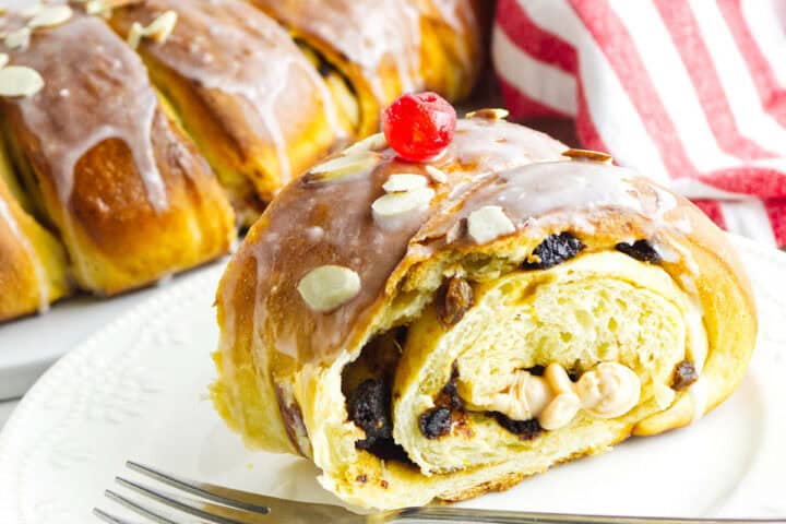 A slice of sweet bread, reminiscent of a festive rosca de reyes, topped with icing, a red cherry, and almond flakes reveals a swirl of raisins and nuts inside. A striped red and white cloth is in the background while a fork rests nearby on a white plate.