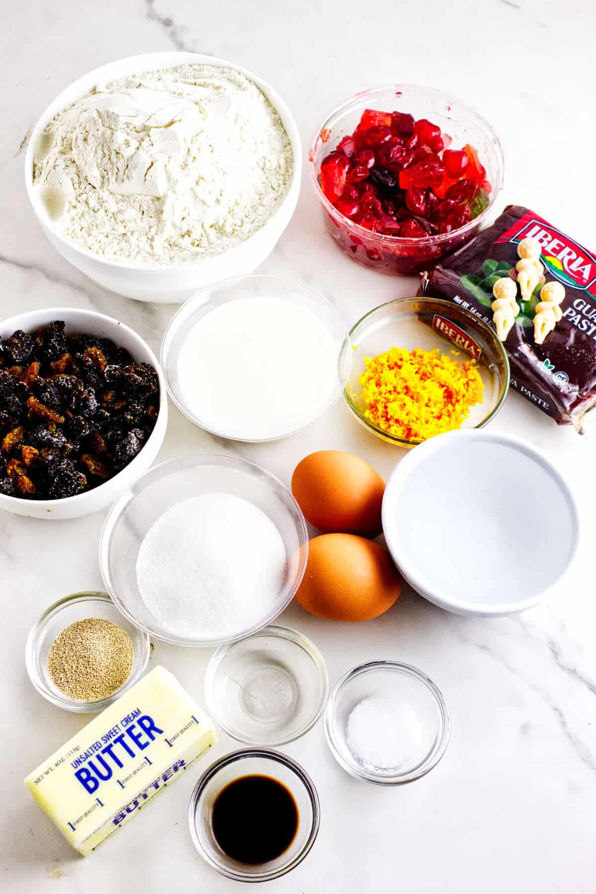 A collection of baking ingredients for a festive rosca de reyes is displayed on a white surface, including flour, cherries, glacé fruits, ginger paste, milk, sugar, butter, eggs, vanilla extract, orange zest, and baking powder.
