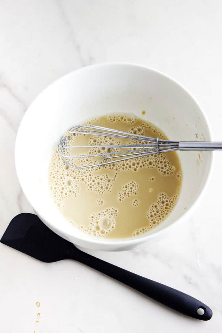 A white bowl filled with a beige, bubbly mixture for a traditional rosca de reyes is accompanied by a metal whisk. A black spatula rests beside the bowl on a light-colored countertop.