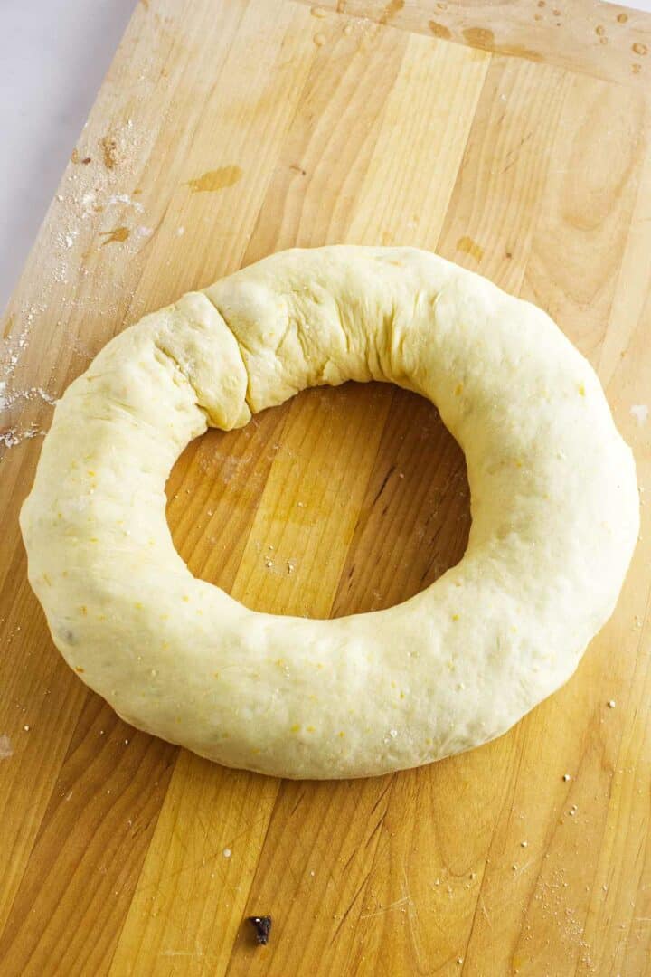 A circular ring of raw dough, reminiscent of a rosca de reyes, rests on a wooden surface. The dough appears lightly floured, with a smooth texture and slight imperfections. The wooden surface shows subtle flour dusting, indicative of a baking preparation process.
