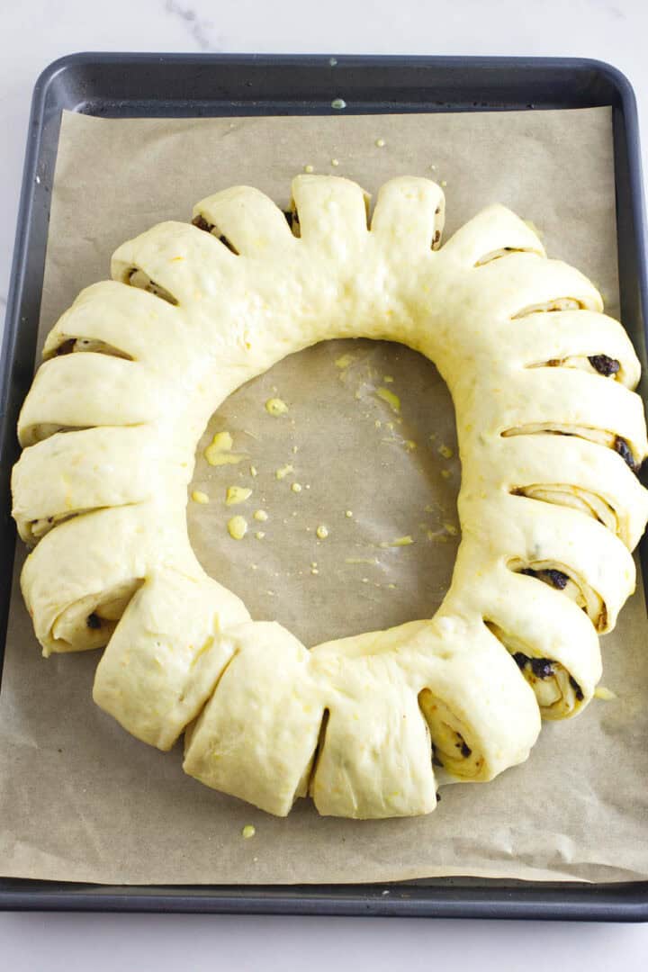 A rosca de reyes, a ring-shaped dough with vertical slits, rests on parchment paper atop a baking tray. Filled with a dark mixture and gently brushed with egg wash, this Wreath of the Kings is primed for baking to golden perfection.