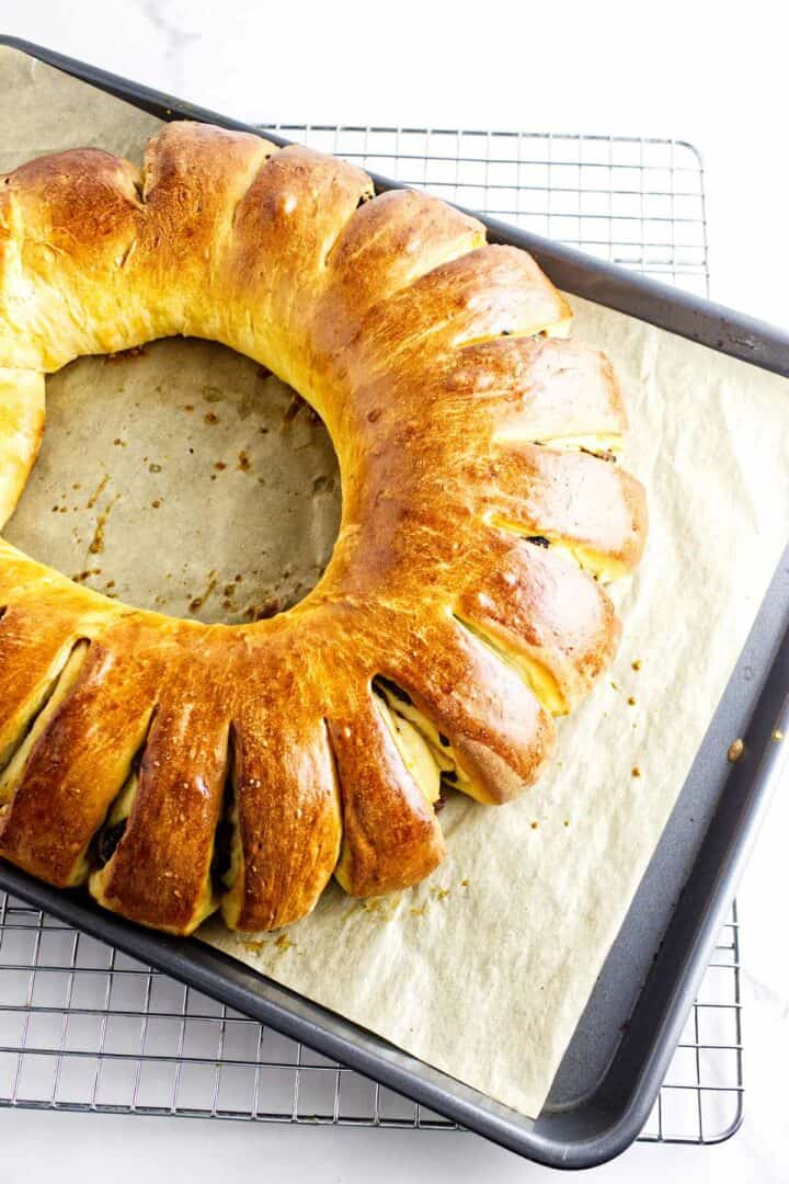 A freshly baked golden-brown rosca de reyes rests on a parchment-lined baking tray, cooling on a wire rack. The bread's curved cuts around the edges hint at its luscious filling, embodying the traditional charm of this Wreath of the Kings.