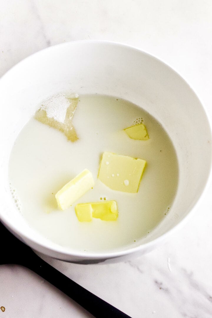 A white bowl containing milk, butter chunks, and sugar sits on a marble countertop, reminiscent of rosca de reyes preparation. A black spoon handle is visible in the foreground.