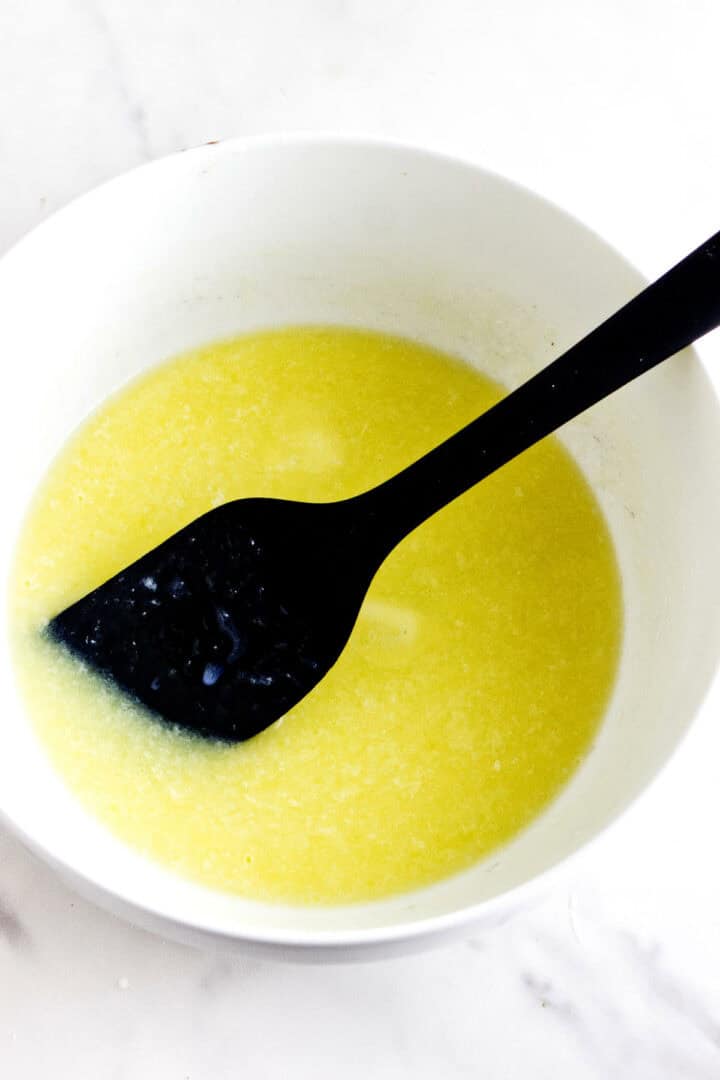 A white bowl containing a mixture reminiscent of the rosca de reyes sits on a white marble surface. Melted butter and sugar combine, cradling a black spatula that's partially submerged in the light yellow liquid, awaiting the next step in creating this festive treat.