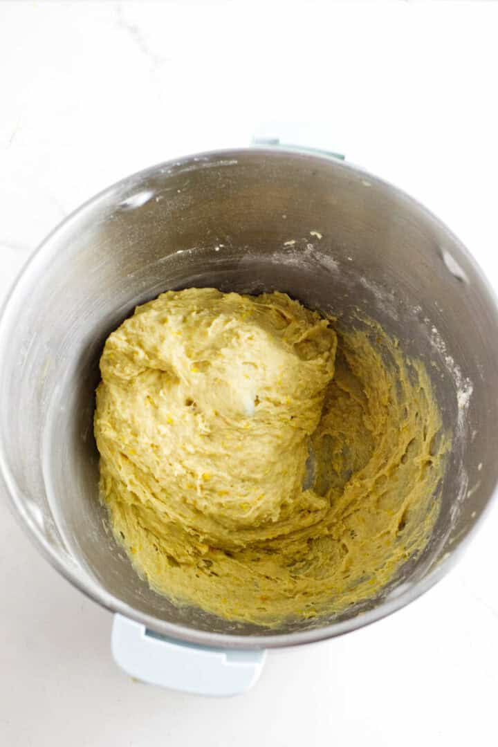 A metal mixing bowl cradles a ball of yellowish, slightly sticky rosca de reyes dough. Its smooth texture and flour-kissed sides promise festive flavor. The bowl rests on a pristine white surface, ready for the next step in creating this traditional treat.