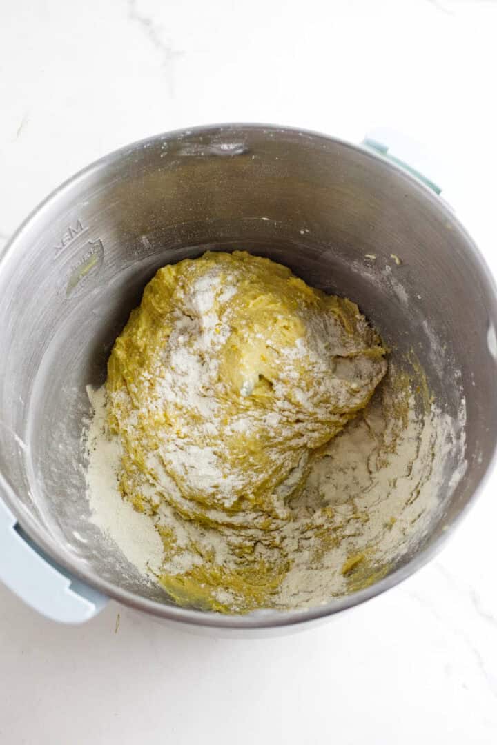 A metallic mixing bowl containing a partially mixed dough for rosca de reyes, with a dusting of flour on top and sides, set against a white background. The dough appears slightly sticky and unformed.