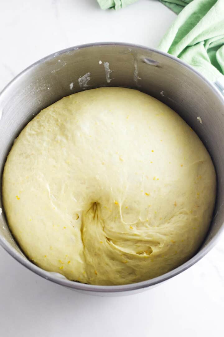 A large bowl of risen dough, reminiscent of a traditional rosca de reyes, is slightly deflated in the center with a smooth and stretchy texture. A green cloth is partially visible in the background, hinting at the festive preparations for Wreath of the Kings celebrations.
