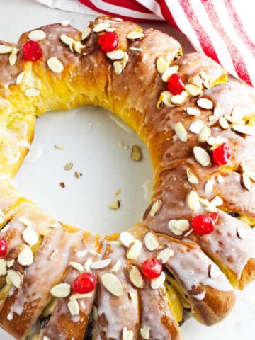 A circular rosca de reyes, or Wreath of the Kings, is topped with white icing, red cherries, and sliced almonds. It rests on a white background with a hint of a red and white striped cloth visible at the top.