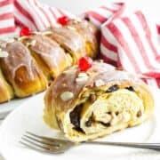 A close-up of a plated slice of rosca de reyes, filled with raisins and topped with almonds and a cherry. In the background, a red and white striped cloth accompanies more of the delicious bread roll. A fork rests invitingly on the plate.
