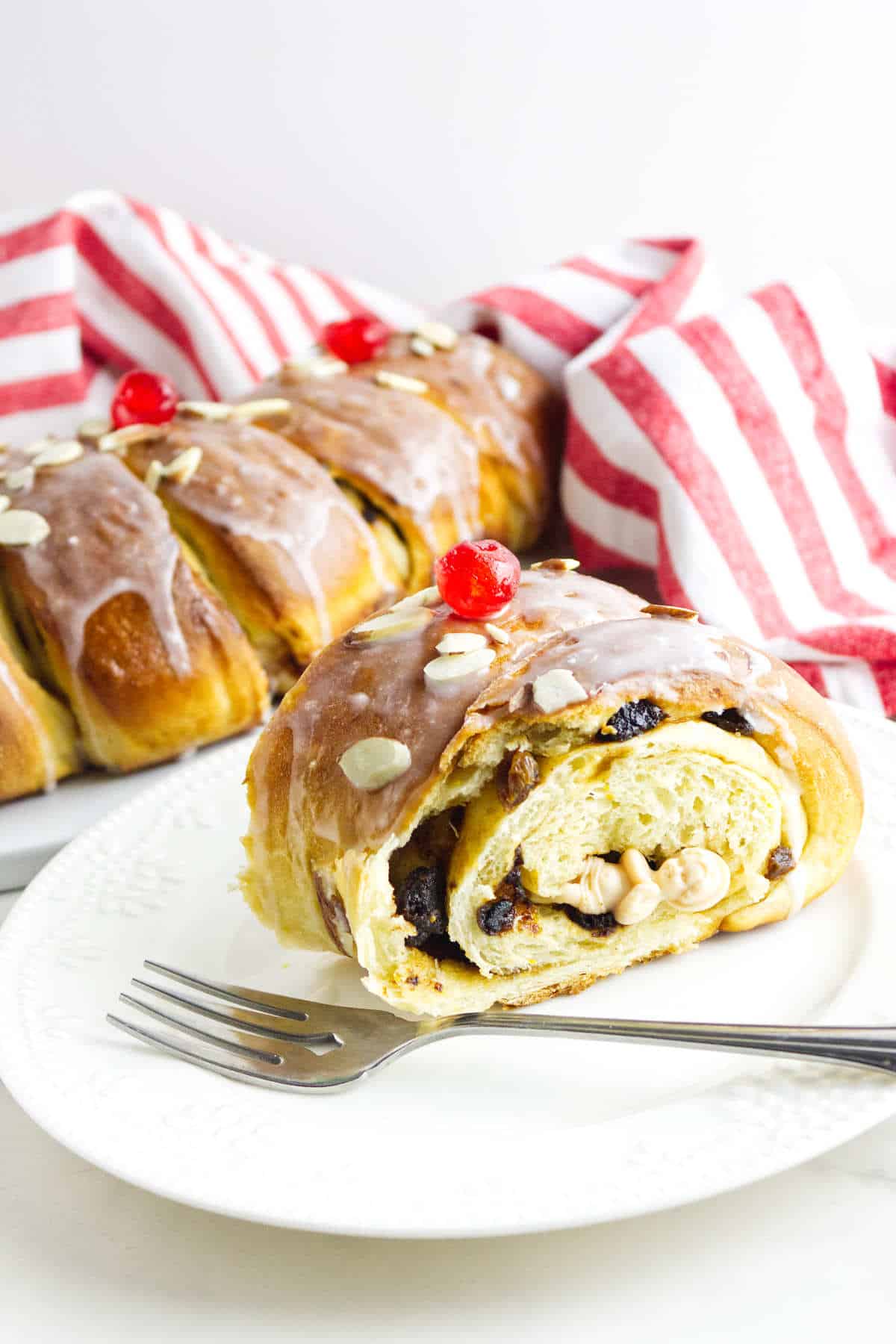 A sliced loaf of sweet bread, reminiscent of a rosca de reyes, with a glazed top garnished with cherries and almond slices, sits on a white plate. A fork rests beside it. A red and white striped cloth is in the background.