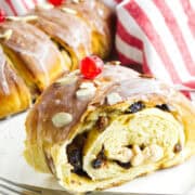 A close-up of a slice of rosca de reyes, this glazed bread roll is topped with a cherry and nuts. The roll features a swirl of raisins inside and is partially sliced. A red and white striped cloth serves as the backdrop, with a fork resting on the plate.