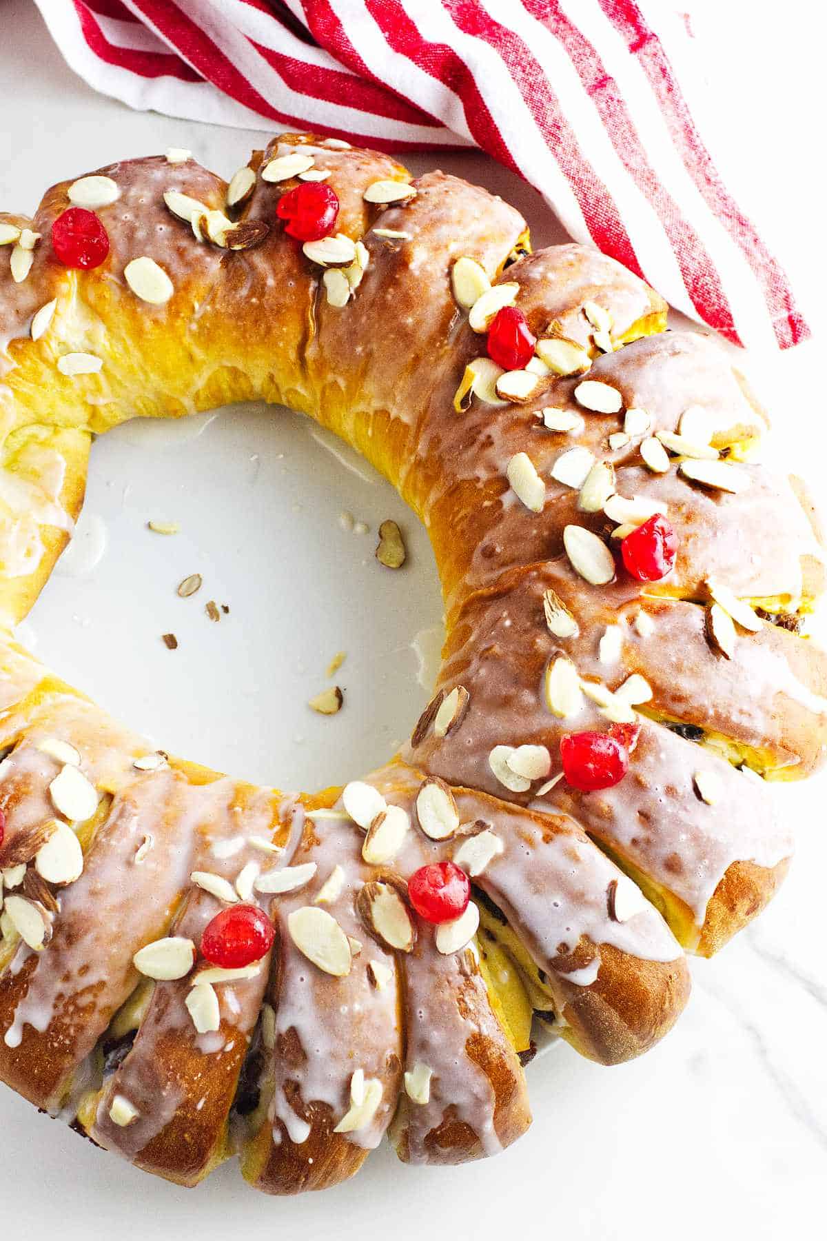 A glazed rosca de reyes, adorned with sliced almonds and cherries, rests elegantly on a white surface. In the background, a red and white striped cloth adds a touch of festive charm to this delightful Wreath of the Kings.