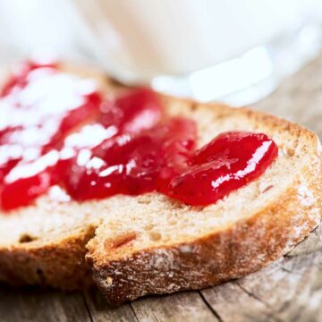 Close-up of a slice of bread with glossy red strawberry jam spread on top, set on a rustic wooden surface. A glass with a white liquid is blurred in the background.