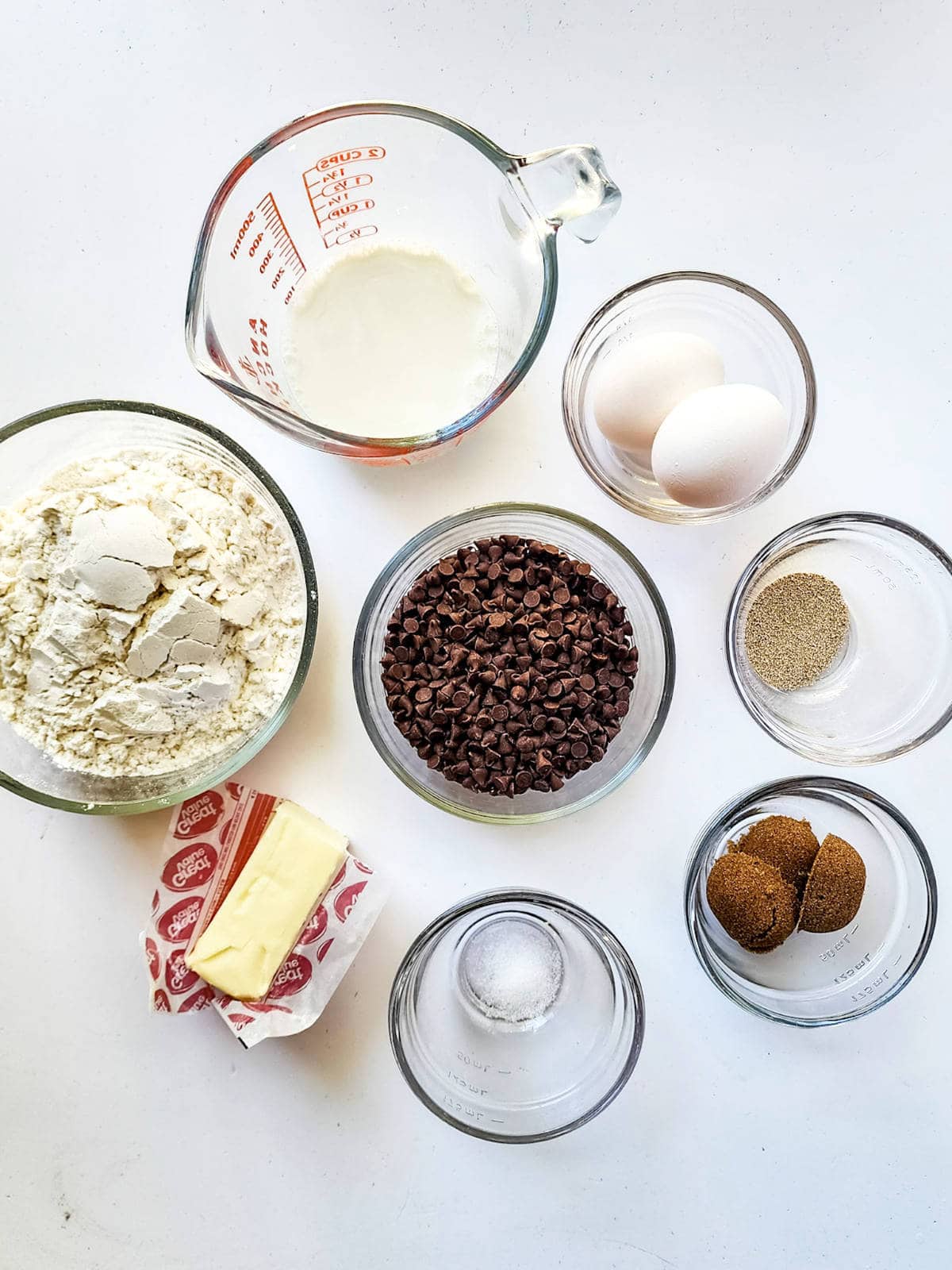 A top view of baking ingredients for a chocolate brioche arranged on a white surface. Includes flour, milk, chocolate chips, eggs, yeast, brown sugar, butter, and salt in separate glass bowls and a measuring cup.