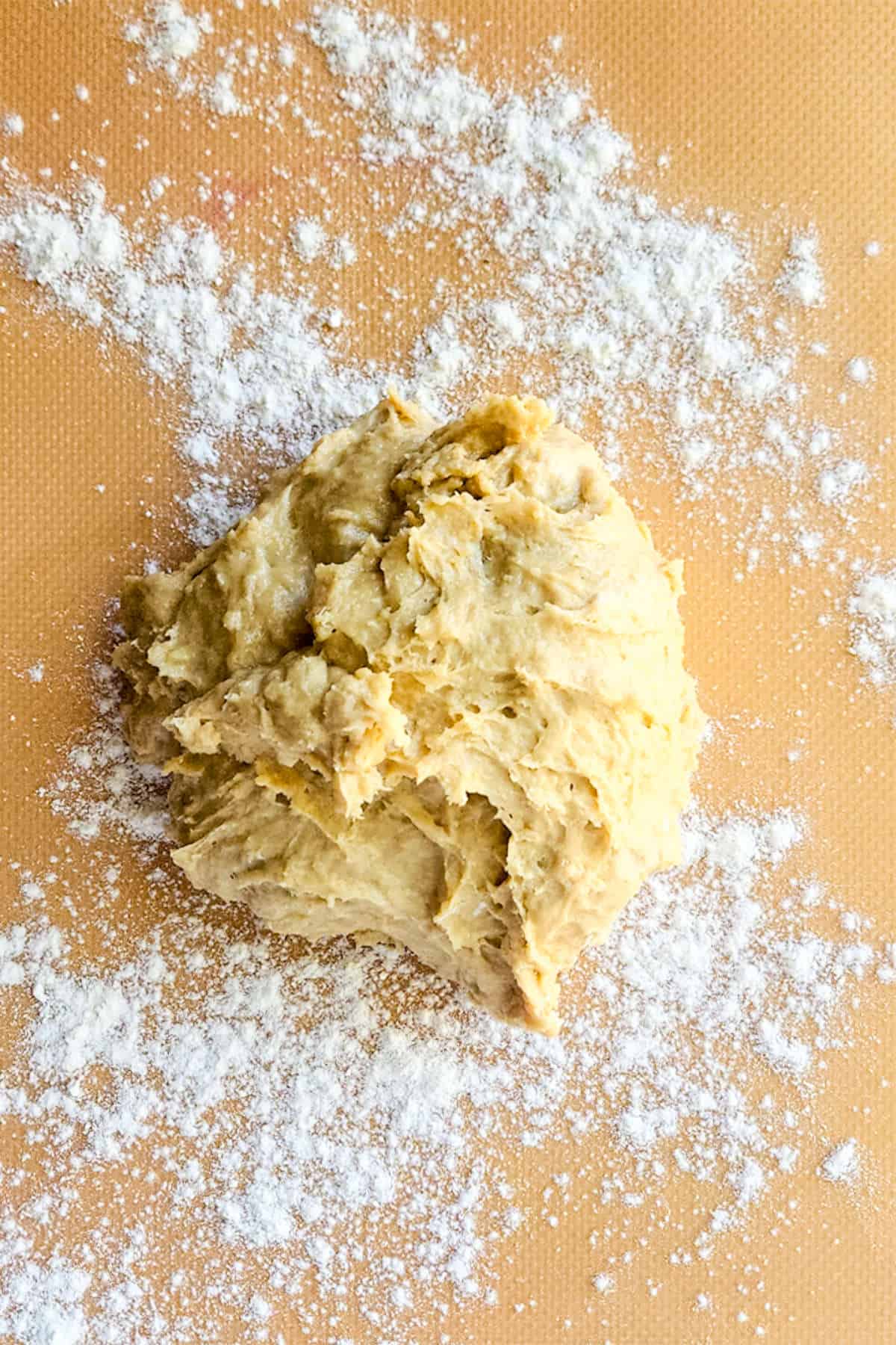 A ball of raw chocolate brioche dough rests on a lightly floured surface, surrounded by scattered flour. The background is a light brown, creating a warm contrast with the pale dough.