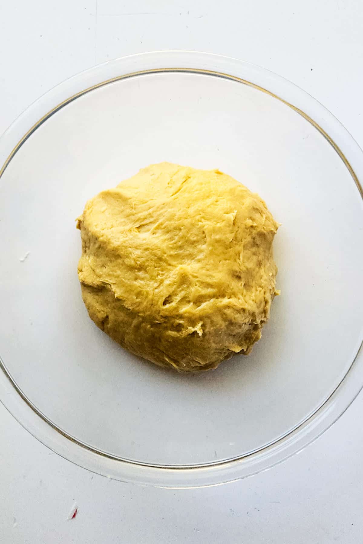 A round brioche boule of dough sits in a transparent glass bowl against a white background. The dough appears smooth and slightly golden, suggesting it is ready for baking or rising.