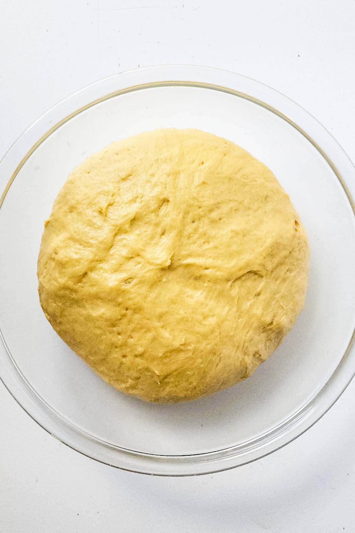 A round ball of raw brioche dough sits in a transparent glass bowl on a white surface. The dough is smooth and evenly formed, ready for baking into a classic brioche boule or further preparation.