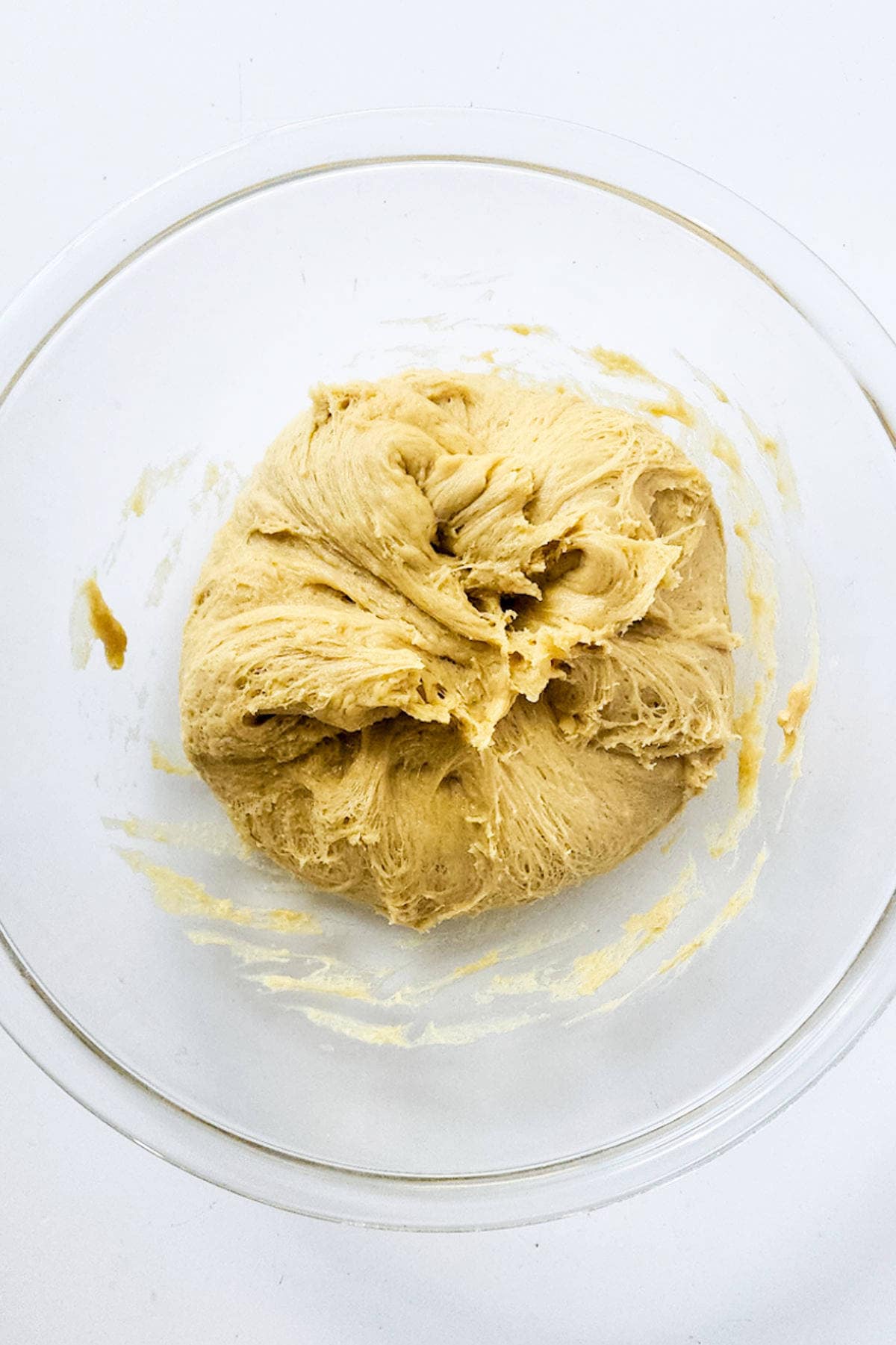 A clear glass bowl contains a ball of raw dough with a slightly rough texture. Resembling a chocolate brioche, the pale yellow dough appears freshly mixed, with remnants clinging to the bowl's sides. It rests elegantly on a white surface.