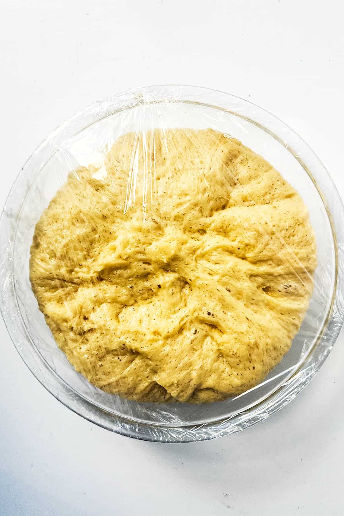 A ball of brioche boule dough is rising in a clear bowl, covered with plastic wrap. The dough has a fluffy texture with visible specks, hinting at spices or grains. It rests serenely on a white surface, embodying the promise of delicate flavors and comforting aromas.