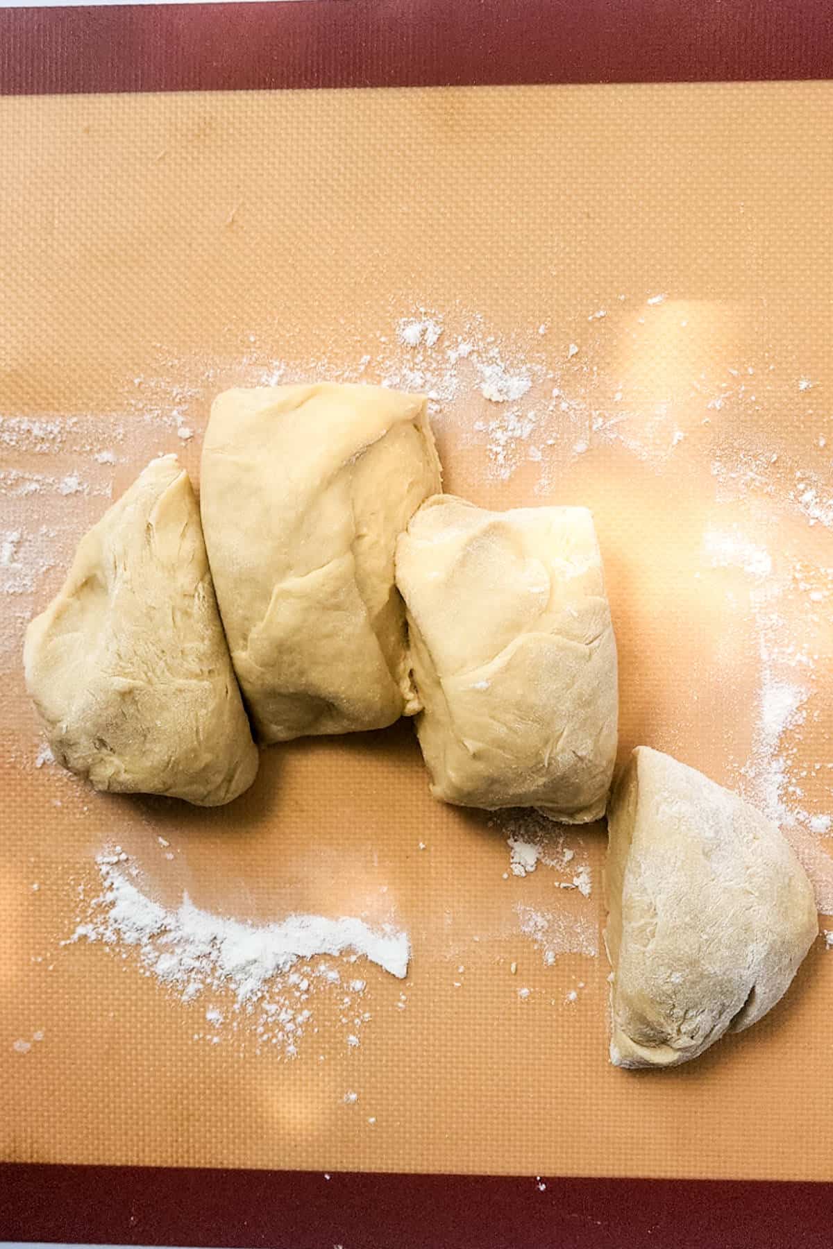 Four pieces of raw dough rest on a lightly floured brown silicone mat, ready for their transformation into delightful treats. Among them, a chocolate brioche boule hints at sweetness to come. The soft-textured dough pieces vary in size and shape, poised for the baking magic ahead.