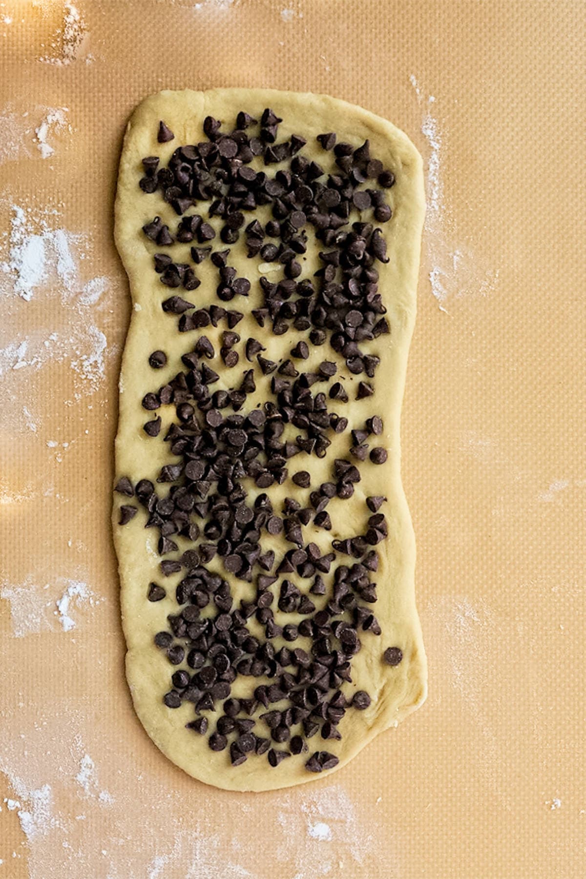 Rolled-out dough, reminiscent of chocolate brioche, is topped with a generous amount of chocolate chips on a lightly floured surface.