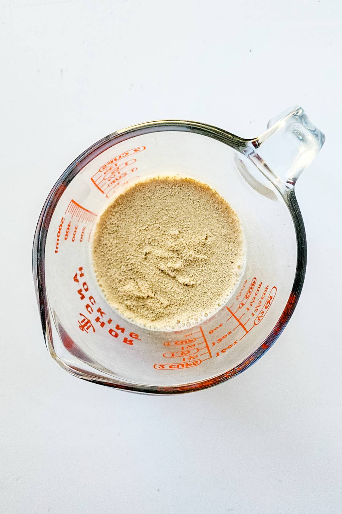 A glass measuring cup, displaying red measurement markings and a handle on the right, holds a light brown powder resembling the warm essence of chocolate brioche. It rests elegantly on a pristine white surface.