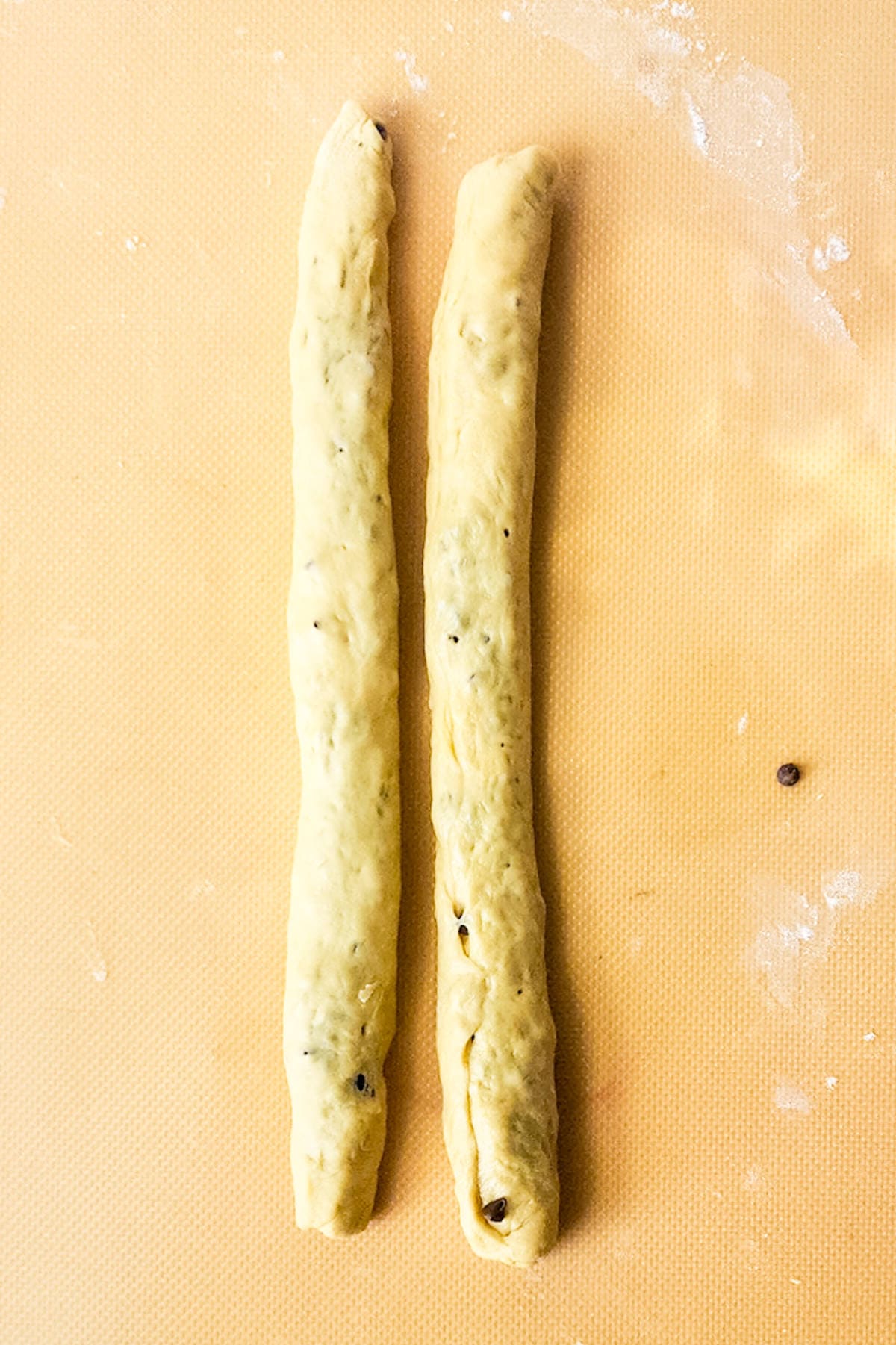 Two elongated pieces of dough with small flecks, reminiscent of chocolate brioche, rest parallel on a textured orange surface, ready for further preparation.