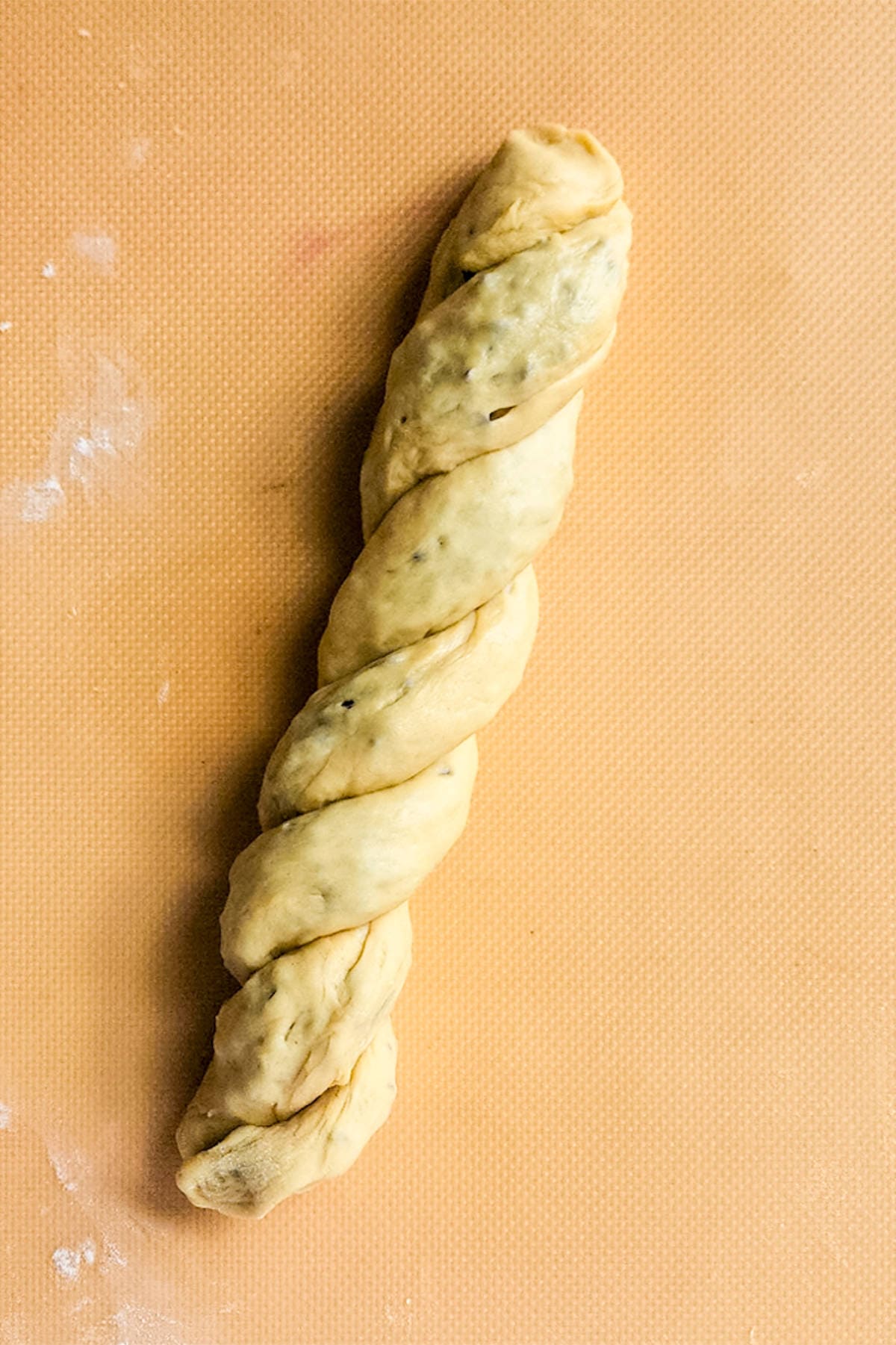 A twisted chocolate brioche dough log rests on an orange surface, likely a baking mat. The dough is evenly twisted, revealing a light, speckled texture that hints at herbs or spices intermingling with rich chocolatey swirls.