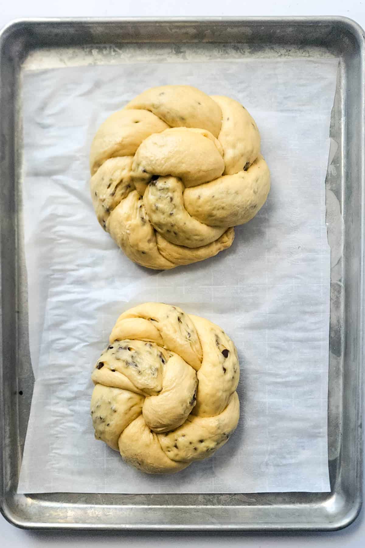 Two braided loaves of uncooked chocolate brioche dough, studded with visible chocolate chips, rest on a parchment-lined baking sheet.