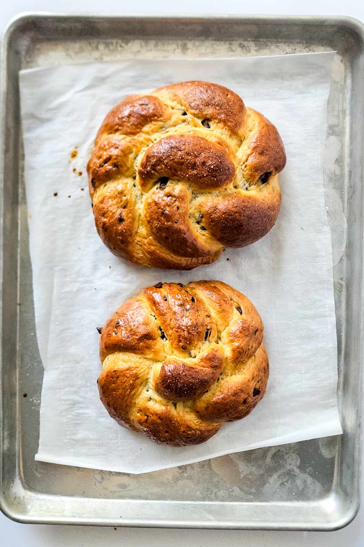 Two freshly baked, braided challah breads sit on a parchment-lined baking sheet alongside a chocolate brioche. The golden-brown loaves have a glossy finish, suggesting a rich texture akin to the soft allure of a classic brioche boule.