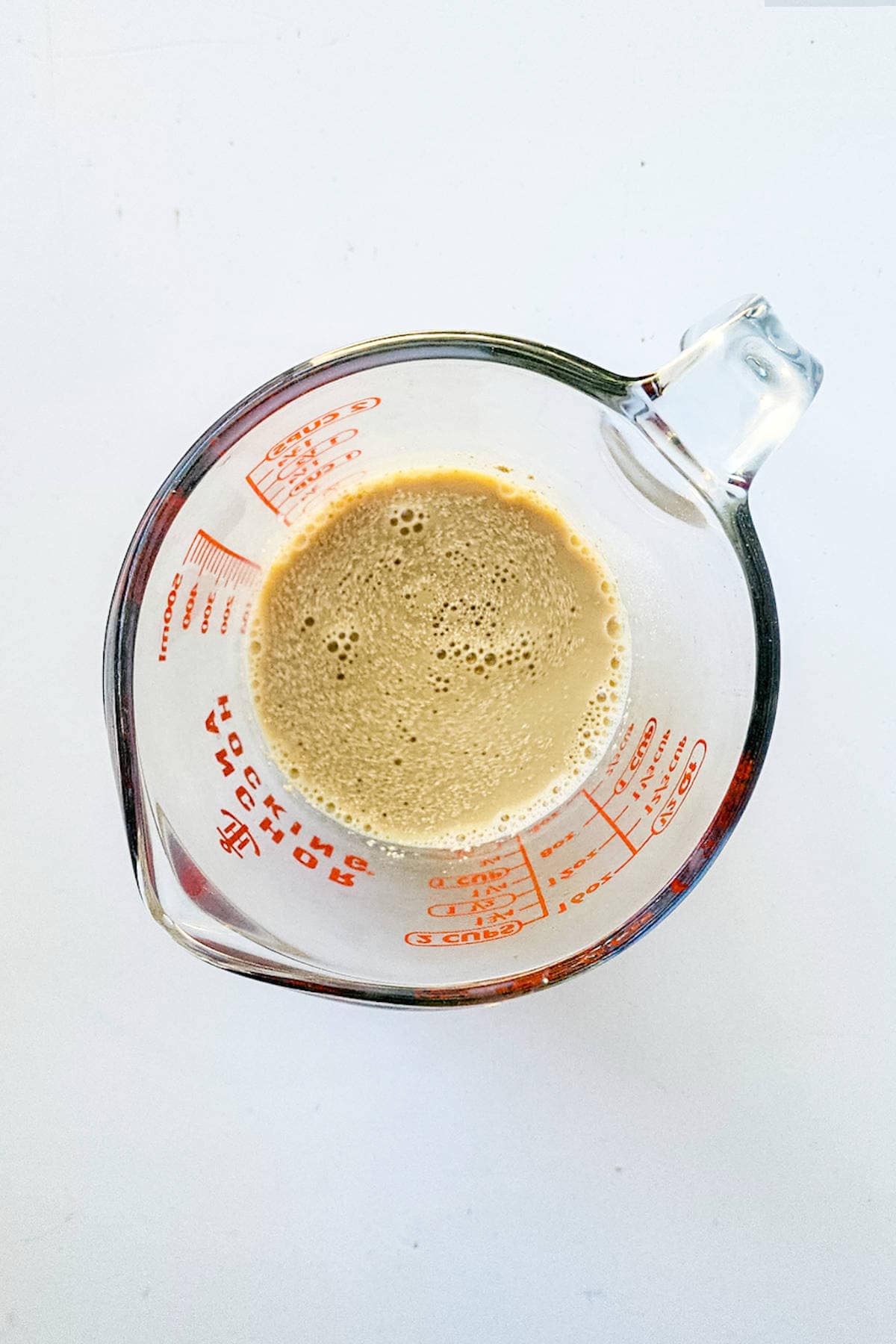 Overhead view of a glass measuring cup containing a frothy brown liquid, reminiscent of melting chocolate brioche. The cup features red measurement markings and is placed on a white surface.