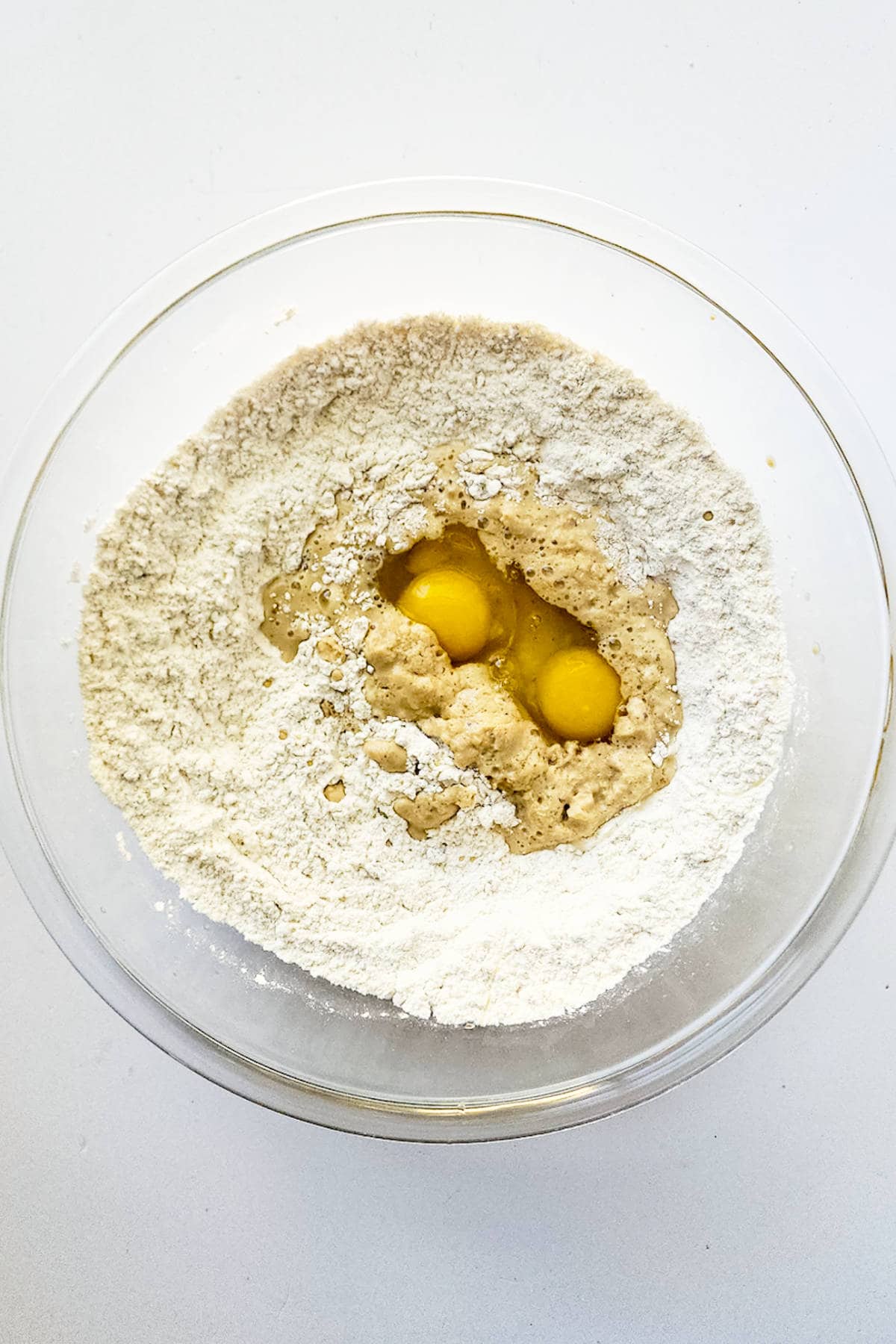 A glass bowl containing flour, poised to create a delightful chocolate brioche, with a well in the center cradling two cracked raw eggs and wet ingredients, ready to be mixed for baking against a white background.