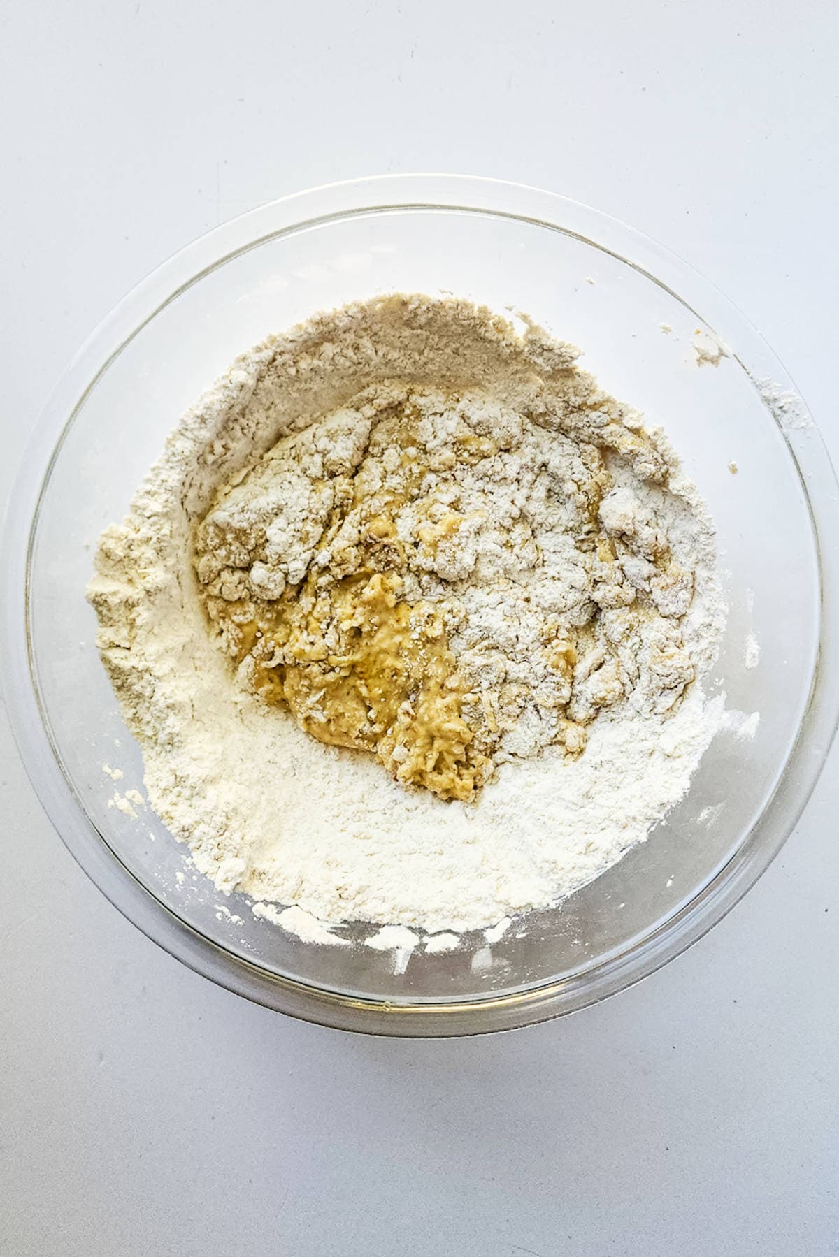 A glass bowl filled with flour and the beginnings of brioche boule dough rests on a white surface. The ingredients are partially combined, hinting at the early stages of crafting this delicious bread.