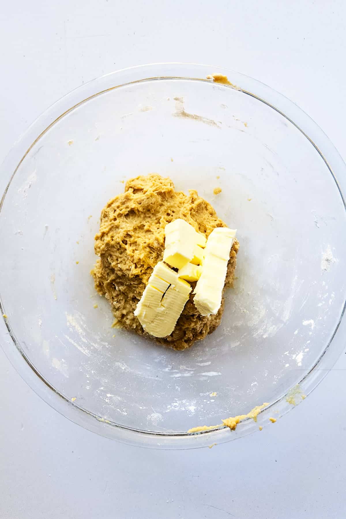 A glass bowl holds a mixture of dough with several cubes of butter on top, evoking the anticipation of crafting a delectable chocolate brioche, set against a plain white background.