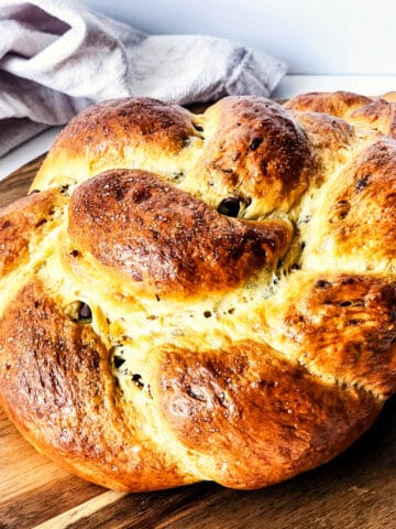 A close-up of a freshly baked round braided brioche boule with a golden-brown crust, placed on a wooden board. A white cloth is partially visible in the background, enhancing the rustic appeal.