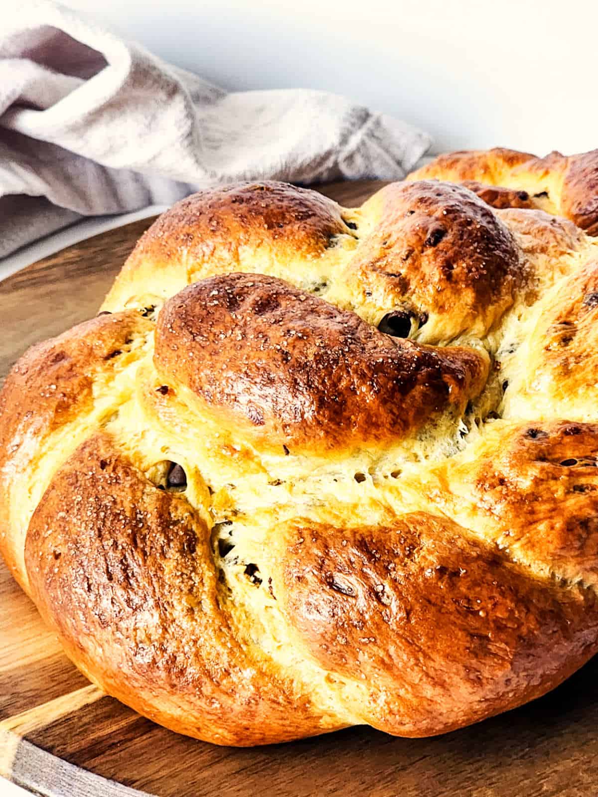 A close-up of a golden brown chocolate brioche loaf, intricately braided and resting on a wooden board. Its glossy finish highlights scattered chocolate chips. A white cloth provides an elegant backdrop.