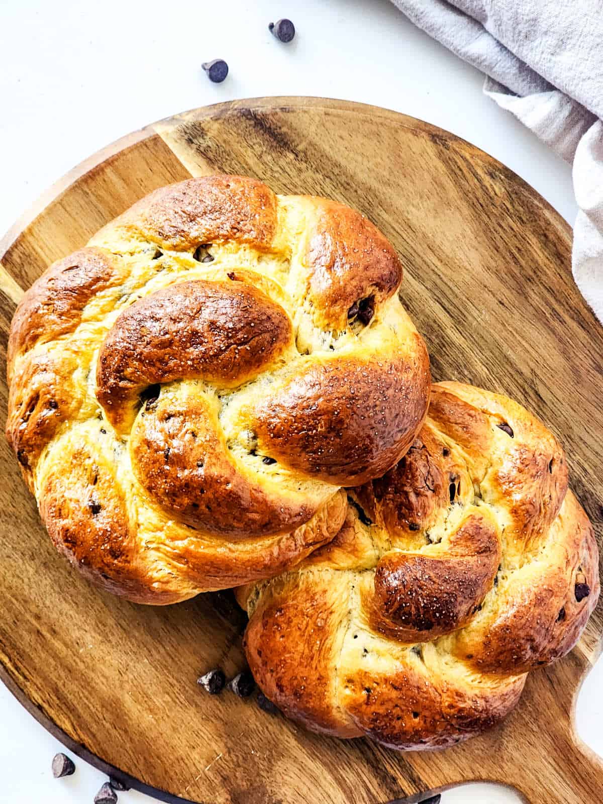 Two freshly baked braided chocolate brioche loaves rest on a round wooden board. The glossy, golden-brown crust gleams enticingly, with scattered chocolate chips nearby. A light-colored cloth is partially visible on the right, complementing this delicious display.