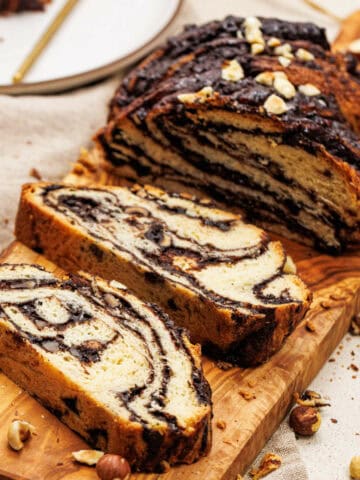 Slices of marble bread with chocolate swirls, reminiscent of a chocolate hazelnut babka, are arranged on a wooden cutting board. Chopped hazelnuts are scattered around the bread. A white plate with more slices is visible in the background.